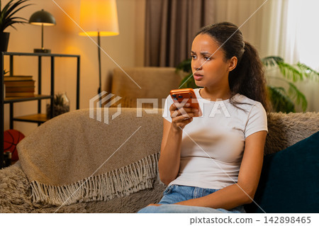 African American young woman on home sofa reads suspicious smartphone message with concern indoors 142898465