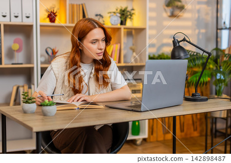 Young woman on home table studying with laptop and book during online lesson writing notes at home 142898487