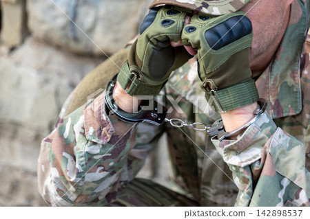 Detained serviceman in camouflage uniform covers his face with handcuffed hands showing emotional tension after capture. Detained serviceman in camouflage uniform covers his face with handcuffed hands showing emotional tension after capture. 142898537