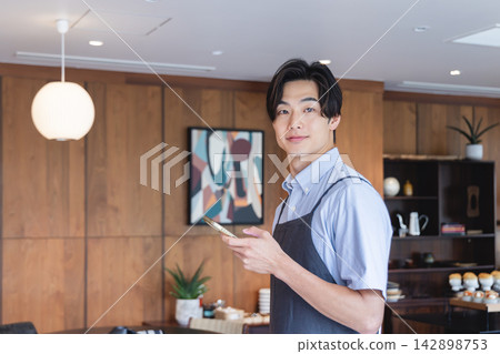 咖啡館裡,一名男性員工拿著智慧型手機。 咖啡館裡,一名男性員工拿著智慧型手機。 142898753