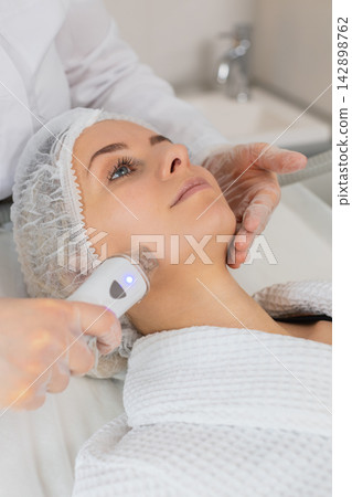 Beautician makes woman RF lifting cosmetology procedure of face and cheek area, collagen mask at spa Beautician makes woman RF lifting cosmetology procedure of face and cheek area, collagen mask at spa 142898762
