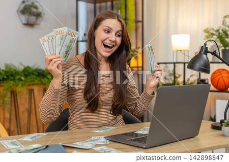 Young woman wins online casino jackpot on laptop celebrates and holds dollar bills as cash on table Young woman wins online casino jackpot on laptop celebrates and holds dollar bills as cash on table 142898847