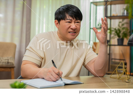 Asian man writing budget notes in notebook, crossing lines, planning big decision under pressure now Asian man writing budget notes in notebook, crossing lines, planning big decision under pressure now 142898848