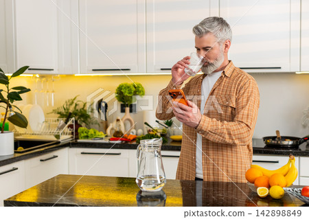 Middle-aged man checks smartphone while drinking water in kitchen for healthy diet hydration routine Middle-aged man checks smartphone while drinking water in kitchen for healthy diet hydration routine 142898849