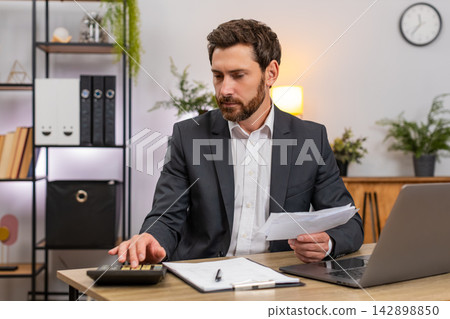 Middle-aged businessman accountant enters numbers on laptop pc checks receipts with calculator signs Middle-aged businessman accountant enters numbers on laptop pc checks receipts with calculator signs 142898850