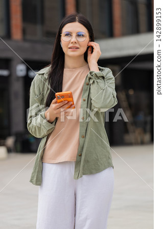 Young woman taking work call walking in city street using smartphone and wireless earbuds outdoors Young woman taking work call walking in city street using smartphone and wireless earbuds outdoors 142899153