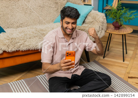 Indian man on home floor celebrating smartphone win with happy smile excited victory gesture indoors Indian man on home floor celebrating smartphone win with happy smile excited victory gesture indoors 142899154