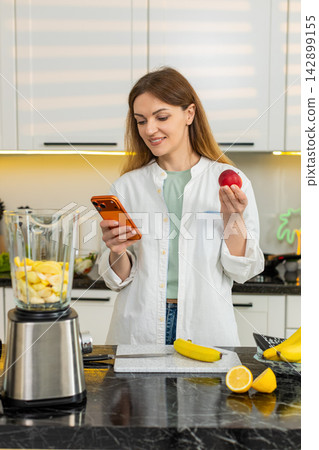 Middle-aged woman choosing fruit for smoothie using smartphone in home kitchen for healthy diet plan 142899155