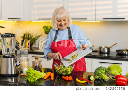 Senior old woman adding cucumber slices to fresh salad in home kitchen for healthy diet meal plans Senior old woman adding cucumber slices to fresh salad in home kitchen for healthy diet meal plans 142899156
