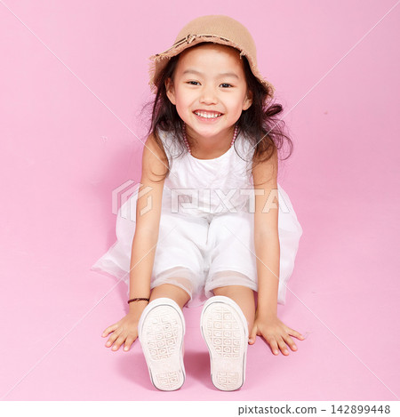 Happy girl in white dress sitting on pink floor with a wicker basket, celebrating childhood joy and fashion. 142899448