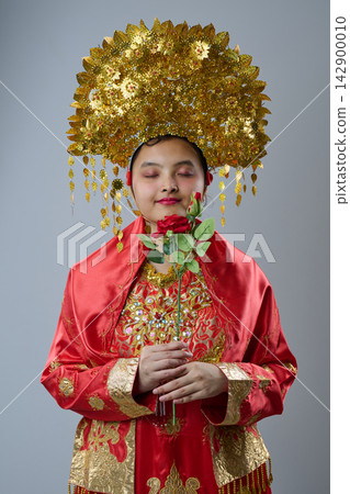 Young Bride In Traditional Red And Gold Costume Holding Rose With Ornate Gold Headdress 142900010