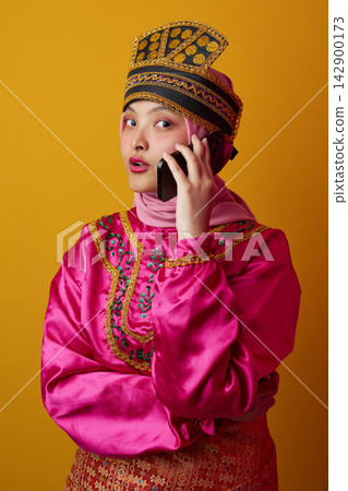 Young Indonesian Asian Woman in Traditional Hijab and Ornate Headdress Talking on Mobile Phone in Colorful Costume Young Indonesian Asian Woman in Traditional Hijab and Ornate Headdress Talking on Mobile Phone in Colorful Costume 142900173