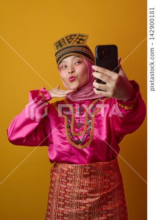 Young Indonesian Asian Woman In Traditional Costume And Hijab Taking Selfie Against Warm Yellow Background 142900181