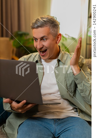 Middle-aged man on home sofa celebrates successful test result on laptop with raised fist and joy Middle-aged man on home sofa celebrates successful test result on laptop with raised fist and joy 142900241
