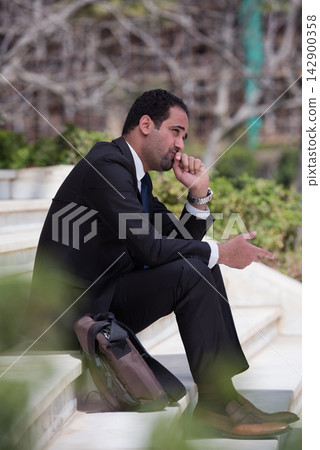 Middle Eastern businessman sitting outdoors in suit and briefcase, thinking during a corporate meeting break Middle Eastern businessman sitting outdoors in suit and briefcase, thinking during a corporate meeting break 142900358