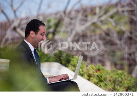 Middle Eastern businessman in suit smiling while working on laptop outdoors in modern startup setting Middle Eastern businessman in suit smiling while working on laptop outdoors in modern startup setting 142900371