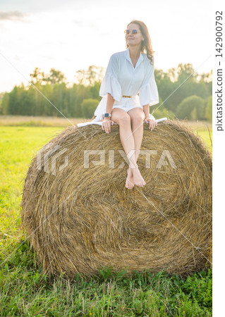 Young woman in white dress sitting barefoot on hay bale in summer countryside. Peaceful rural lifestyle represents slow living, freedom and relaxation in nature Young woman in white dress sitting barefoot on hay bale in summer countryside. Peaceful rural lifestyle represents slow living, freedom and relaxation in nature 142900792