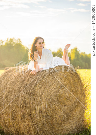 Young woman in white dress lying barefoot on hay bale at sunset. Carefree summer moment represents slow living, relaxation and freedom in countryside Young woman in white dress lying barefoot on hay bale at sunset. Carefree summer moment represents slow living, relaxation and freedom in countryside 142900805
