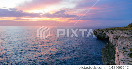 The morning light casts a soft glow across the calm sea surface while dramatic rock formations stand tall against the vibrant colorful horizon The morning light casts a soft glow across the calm sea surface while dramatic rock formations stand tall against the vibrant colorful horizon 142900942