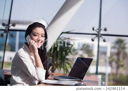 Young businesswoman smiling on phone at laptop in a modern open office meeting space Young businesswoman smiling on phone at laptop in a modern open office meeting space 142901114