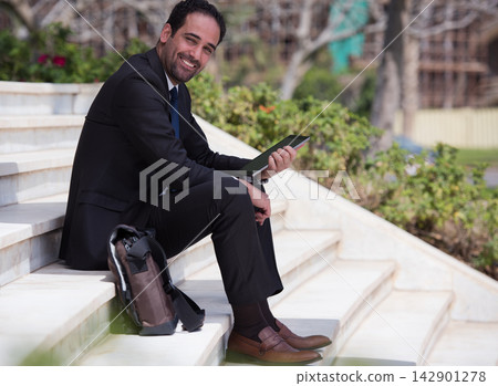 Middle Eastern businessman sitting on outdoor steps with tablet smiling in a modern corporate setting Middle Eastern businessman sitting on outdoor steps with tablet smiling in a modern corporate setting 142901278