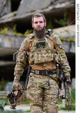 Bearded Soldier In Tactical Camouflage Gear Holding Helmet And Rifle In Urban Environment Bearded Soldier In Tactical Camouflage Gear Holding Helmet And Rifle In Urban Environment 142901299