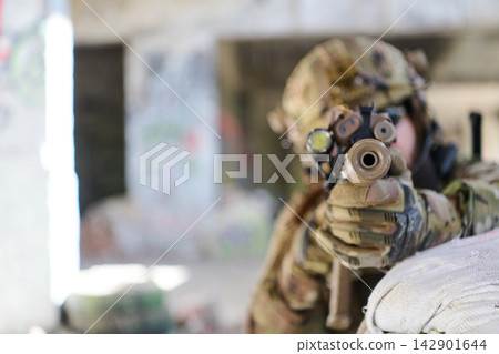 Soldier in Camouflage Aiming Rifle Through Scope From Behind Sandbag in Urban Ruins Soldier in Camouflage Aiming Rifle Through Scope From Behind Sandbag in Urban Ruins 142901644