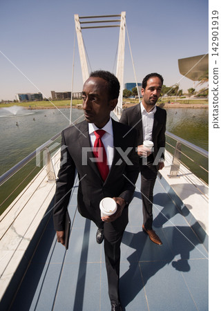 Two businessmen walking on a pedestrian bridge with coffee and briefcase near an urban lake Two businessmen walking on a pedestrian bridge with coffee and briefcase near an urban lake 142901919