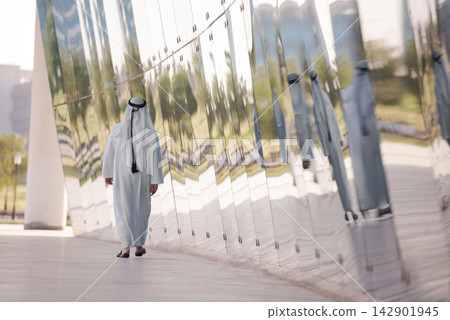Man in white thobe walking along a reflective curved wall with repeated mirrored reflections Man in white thobe walking along a reflective curved wall with repeated mirrored reflections 142901945