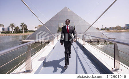 Businessman walking confidently across modern bridge toward glass pyramid office building on waterfront Businessman walking confidently across modern bridge toward glass pyramid office building on waterfront 142901946