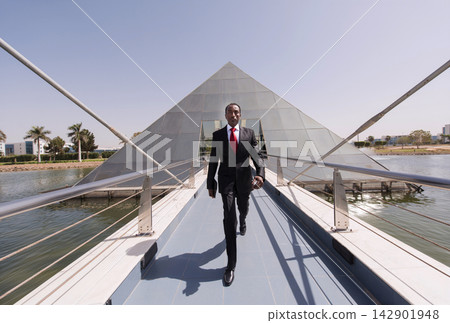 Business executive walking confidently on a bridge toward a modern glass pyramid building over water Business executive walking confidently on a bridge toward a modern glass pyramid building over water 142901948