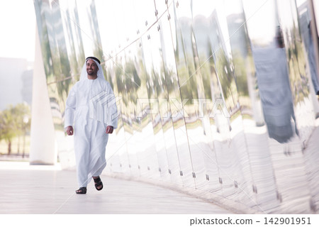 Smiling Arab man in traditional dress walking by reflective modern architecture in an urban park Smiling Arab man in traditional dress walking by reflective modern architecture in an urban park 142901951