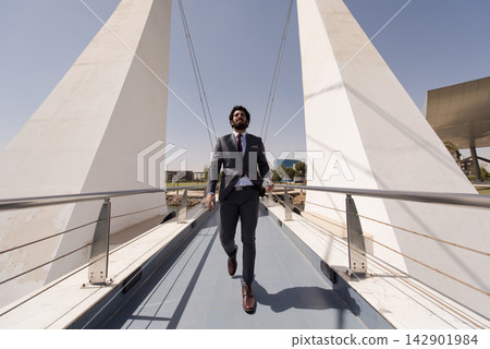 Businessman walking confidently across modern pedestrian bridge in suit and tie on sunny day Businessman walking confidently across modern pedestrian bridge in suit and tie on sunny day 142901984