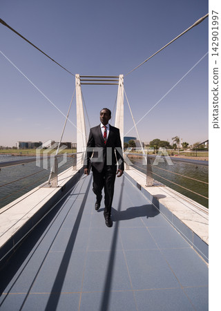 Confident businessman walking across modern pedestrian bridge in urban waterfront business district Confident businessman walking across modern pedestrian bridge in urban waterfront business district 142901997