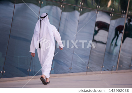 Man in traditional white kandura walking past reflective glass facade of modern building Man in traditional white kandura walking past reflective glass facade of modern building 142902088