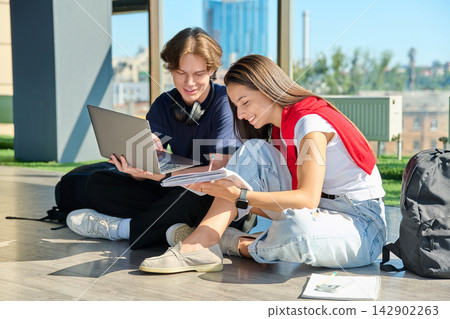 Two young students studying with laptop and notebook sitting on floor near big window Two young students studying with laptop and notebook sitting on floor near big window 142902263