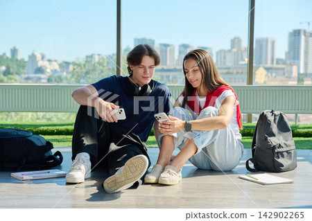 Two young students sitting on floor and looking at smartphones near window Two young students sitting on floor and looking at smartphones near window 142902265