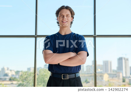 Smiling young man standing with crossed arms near large window with city view 142902274