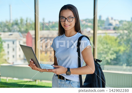 Smiling female student wearing eyeglasses holding laptop with city window background 142902280