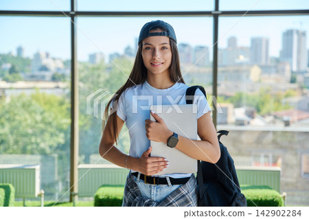 Smiling female student in cap holding laptop computer against city window Smiling female student in cap holding laptop computer against city window 142902284