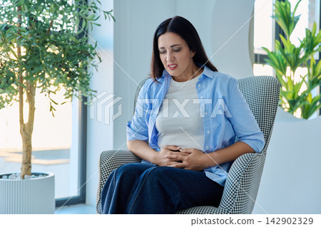 Middle aged woman holding stomach suffering from abdominal pain in armchair Middle aged woman holding stomach suffering from abdominal pain in armchair 142902329