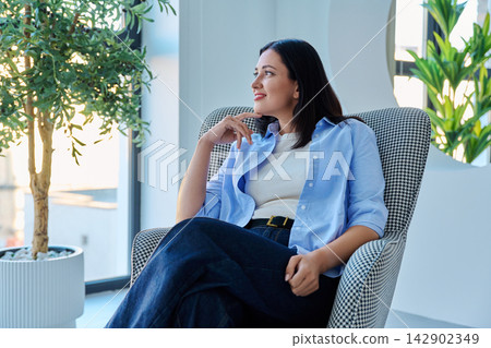 Charming middle aged woman sitting in armchair smiling thoughtfully 142902349