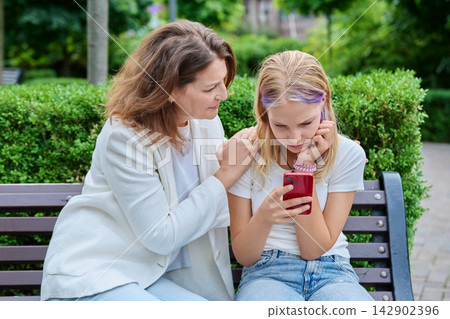 Caring mother embracing teenage daughter using smartphone sitting on bench Caring mother embracing teenage daughter using smartphone sitting on bench 142902396