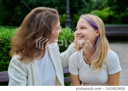 Caring mother touching cheek of smiling teenage daughter sitting on bench Caring mother touching cheek of smiling teenage daughter sitting on bench 142902400