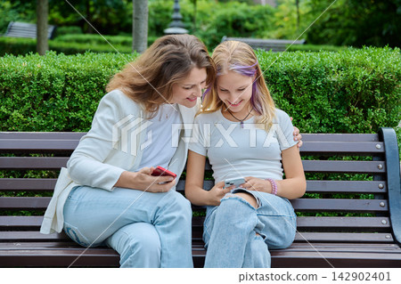Mother leaning towards teenage daughter using smartphone on bench in park Mother leaning towards teenage daughter using smartphone on bench in park 142902401
