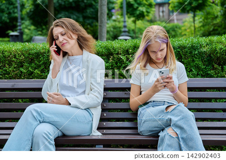 Mother talking on phone and daughter looking at smartphone on bench park Mother talking on phone and daughter looking at smartphone on bench park 142902403