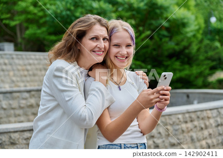 Smiling mother and daughter posing with smartphone looking at camera park Smiling mother and daughter posing with smartphone looking at camera park 142902411