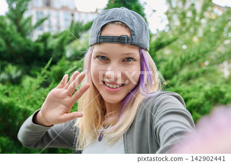Smiling teenage girl with purple hair strands waving hand taking selfie outdoors Smiling teenage girl with purple hair strands waving hand taking selfie outdoors 142902441