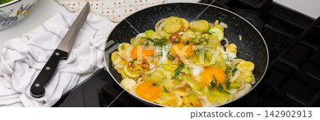 Fried zucchini is cooked with scrambled eggs in a pan on a stovetop. The scene shows a kitchen preparing a meal. Fresh ingredients are visible, creating a simple dish, banner Fried zucchini is cooked with scrambled eggs in a pan on a stovetop. The scene shows a kitchen preparing a meal. Fresh ingredients are visible, creating a simple dish, banner 142902913