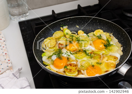 Fried zucchini is cooked with scrambled eggs in a pan on a stovetop. The scene shows a kitchen preparing a meal. Fresh ingredients are visible, creating a simple dish Fried zucchini is cooked with scrambled eggs in a pan on a stovetop. The scene shows a kitchen preparing a meal. Fresh ingredients are visible, creating a simple dish 142902946